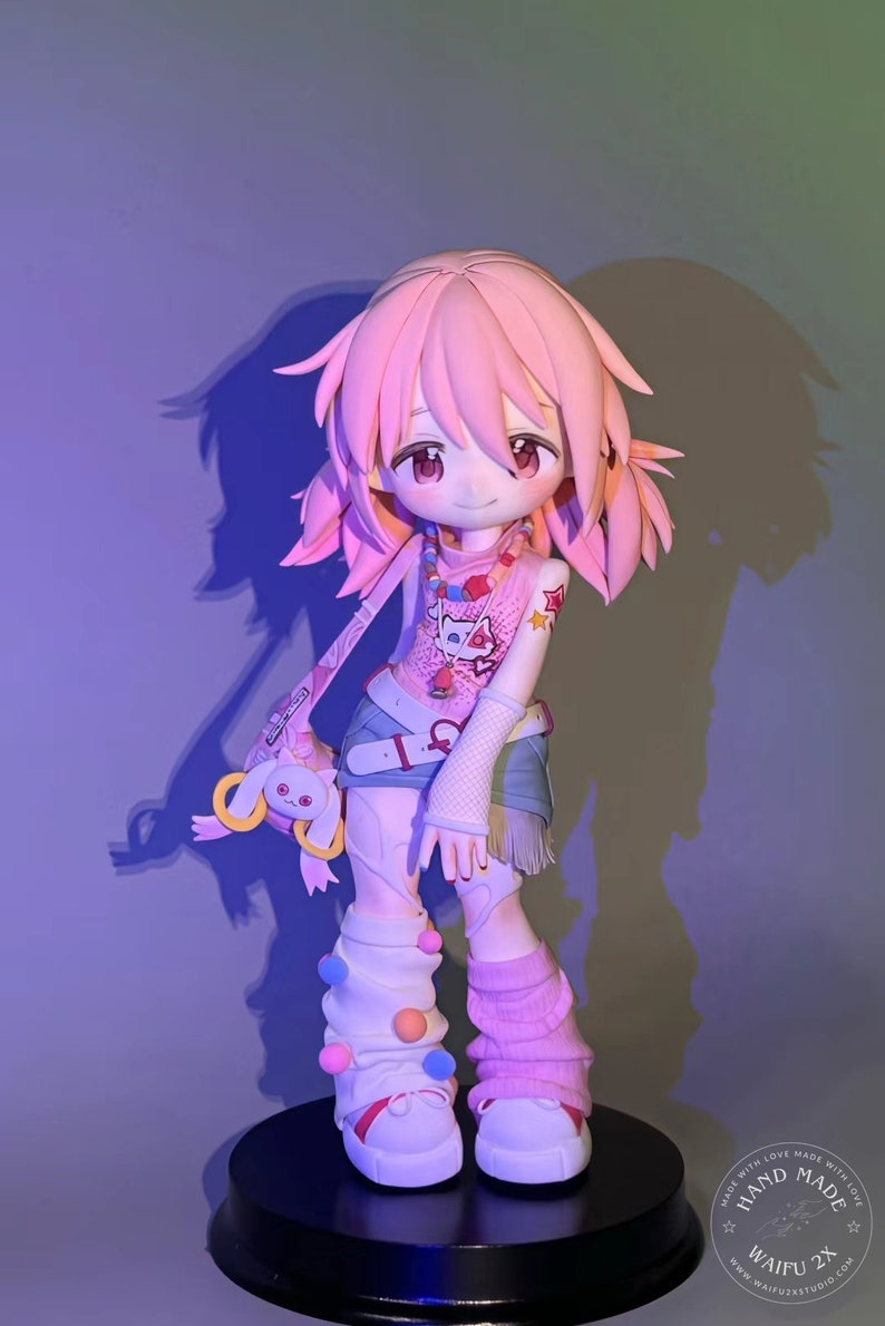 May include: A pink and white anime figurine with pink hair, wearing a pink and white shirt with a cat design, blue denim shorts, and pink and white socks with colorful pom-poms. The figurine is holding a pink and white plush toy. The figurine is standing on a black base with a white sticker that says "Hand Made with Love Made Without Love" and "Waifu 2X".