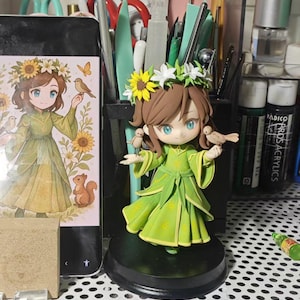 以下が含まれることがあります： 緑色のドレスを着て、花の冠をかぶり、小さな鳥を2羽持った少女の手作りの置物。置物は黒い台座の上にあり、近くの携帯電話の画面には同様のイラストが描かれています。