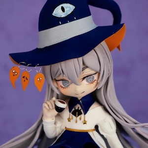 May include: A handcrafted figurine of a witch with long gray hair, wearing a navy blue hat with an eye design and orange accents. The witch is dressed in a white and navy blue dress and holding a tiny cup. Three orange ghost earrings dangle from the hat.