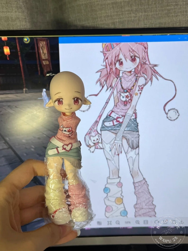 May include: A small, light pink and white doll with long pink hair and a pink and white outfit. The doll is holding a small white bag with a pink bow. The doll is standing on a white surface. The doll is a handmade doll.