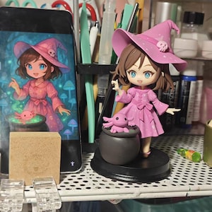以下が含まれることがあります： ピンク色の魔女のフィギュア。お揃いの帽子とドレスを着て、小さなウーパールーパーを持っています。フィギュアは黒い台座の上にあり、同様の画像を表示する電話の隣にあります。背景には画材と棚があります。