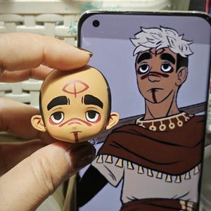 May include: A small, hand-painted figurine of a bald-headed character with red markings on the forehead, eyes, and chin. The figurine is held in a hand, with a smartphone displaying a matching cartoon character in the background. The cartoon character has white hair and similar facial markings.