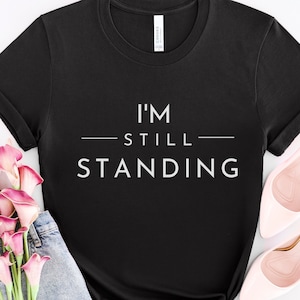 May include: Black t-shirt with white text that reads "I'm Still Standing".