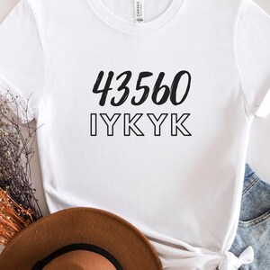 以下が含まれることがあります： 黒い文字で「43560 IYKYK」と書かれた白いTシャツ。