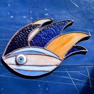 May include: A stained glass fish with a blue, orange, and white design. The fish has a large, round eye.