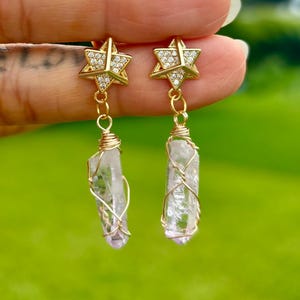 May include: A pair of gold-toned earrings featuring a six-pointed star stud encrusted with small clear stones. A dangling, raw, clear crystal point is secured to each star by gold-colored wire wrapping, creating a drop-style earring design.