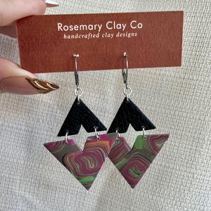 May include: A pair of handcrafted clay earrings. The earrings feature a black triangle at the top and a marbled triangle in shades of pink, green, and gray below. The earrings are suspended from silver-colored ear wires. The tag reads "Rosemary Clay Co handcrafted clay designs."