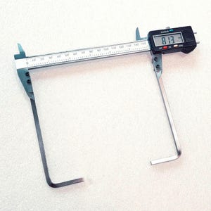 May include: A digital calliper with long, L-shaped metal arms for measuring internal or external dimensions. The device features a silver metal scale marked in millimetres and inches, and a black digital display showing the reading 81.3 mm.