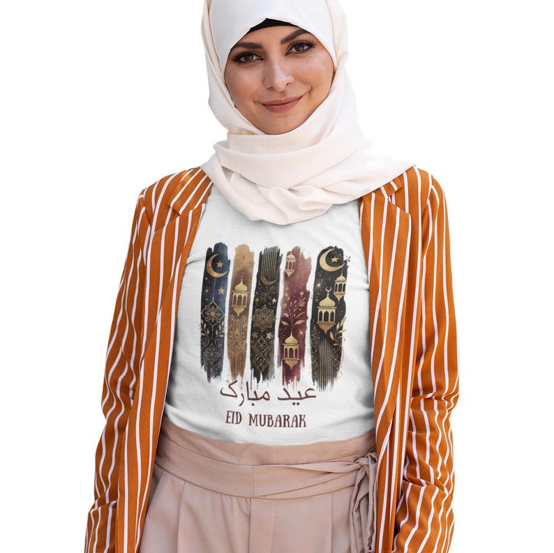 May include: White t-shirt with a colourful graphic design featuring Islamic patterns, lanterns, and crescent moons. The text "Eid Mubarak" is written in Arabic and English.