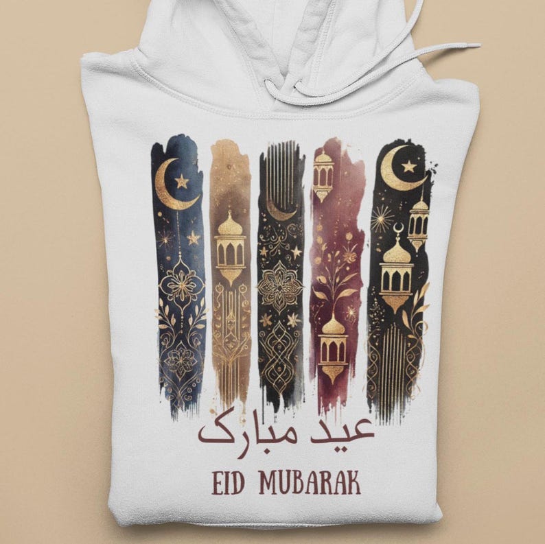 May include: White hoodie featuring a gold and black design of crescent moons, stars, and lanterns. The text "Eid Mubarak" is written in Arabic and English.