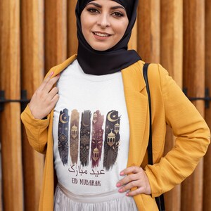 May include: White t-shirt with a gold and black graphic design featuring a crescent moon, stars, and lanterns. The text "Eid Mubarak" is written in Arabic and English.