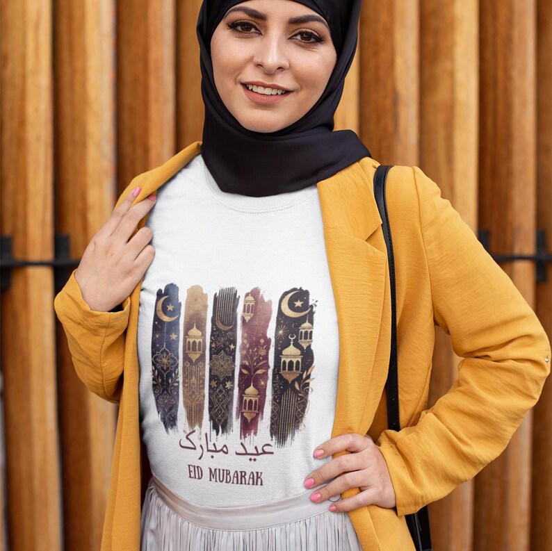 May include: White t-shirt with a gold and black graphic design featuring a crescent moon, stars, and lanterns. The text "Eid Mubarak" is written in Arabic and English.