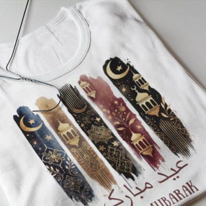 May include: White t-shirt with a gold and black design featuring crescent moons, stars, and lanterns. The text "Eid Mubarak" is written in Arabic script.