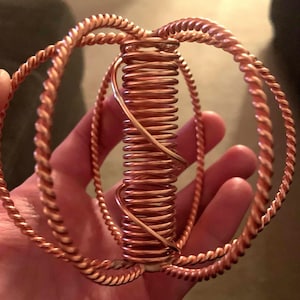 Può includere: Una scultura fatta a mano color rame tenuta in una mano. La scultura presenta una spirale centrale, due anelli ritorti e un elemento decorativo in filo di ferro. Il pezzo ha una calda lucentezza metallica e sembra essere realizzato in filo di rame.