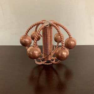 Puede incluir: Una escultura de cobre hecha a mano con una bobina cilíndrica central, rodeada por seis esferas de cobre. Las esferas están conectadas a la bobina central por alambres de cobre torcidos y curvados. La escultura se exhibe sobre una superficie marrón oscuro.