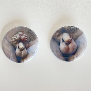 May include: Two round, white ceramic buttons featuring a detailed image of a sculpture of a person with their arms crossed over their chest. The sculpture is in a light brown colour.