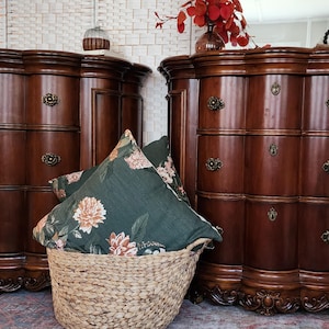 May include: Two antique wooden dressers with ornate details and brass hardware. A woven basket holds floral pillows in front of the dressers. The dressers are a rich, dark brown color, and the scene is set on a patterned rug.