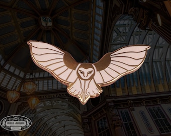 May include: A gold and white enamel pin featuring a barn owl with its wings spread. The owl is flying with its head turned to the side. The pin is a decorative accessory.
