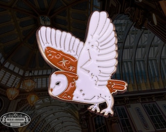May include: A white enamel pin with a brown owl design. The owl has a white body with brown wings and a brown face. The owl has a star pattern on its wings. The pin is a great addition to any owl lover's collection.