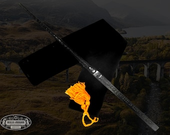 May include: A black wand with a dark wood finish and a black velvet pouch with a gold tassel. The wand is lying on a black surface with a blurred background of a landscape.