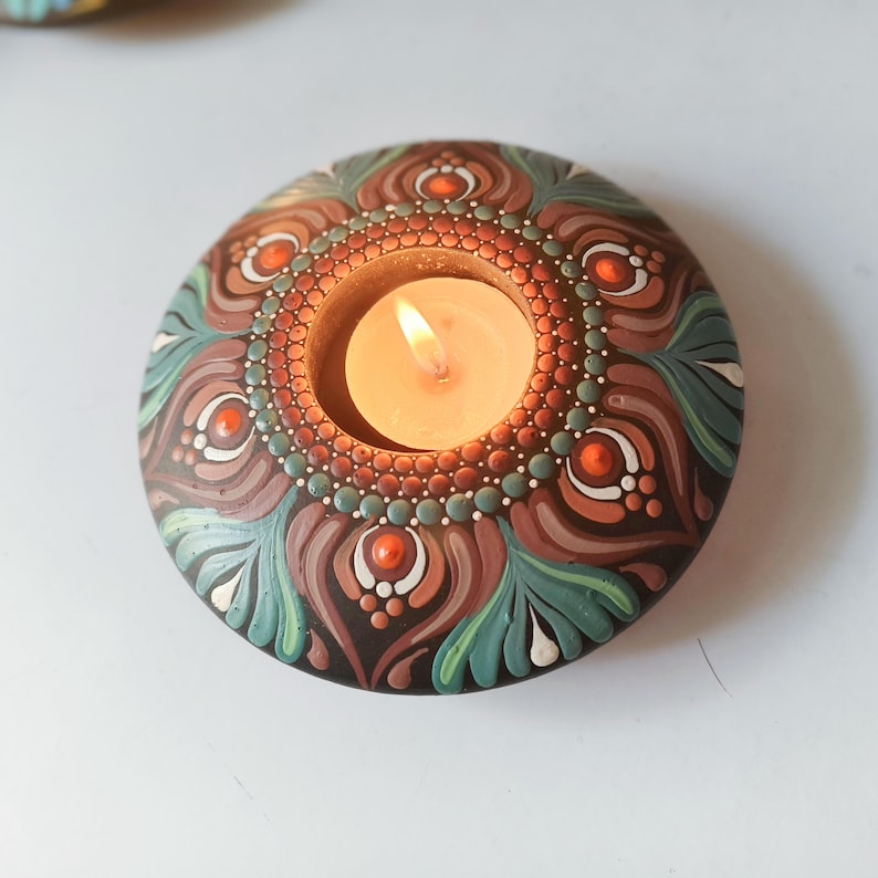 K&ouml;nnte beinhalten: Ein runder, handbemalter Kerzenhalter mit einer brennenden Kerze. Der Halter hat ein detailliertes Mandala-Design in Braun-, Gr&uuml;n-, Orange- und Wei&szlig;t&ouml;nen. Die Kerzenflamme leuchtet in der Mitte und erzeugt eine warme Atmosph&auml;re.