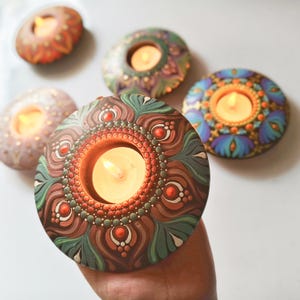 K&ouml;nnte beinhalten: Handbemalte, runde Kerzenhalter mit aufwendigen Mandala-Designs. Die Halter haben eine zentrale Aussparung f&uuml;r ein Teelicht, umgeben von detaillierten Mustern in Braun-, Gr&uuml;n-, Rot- und Blaut&ouml;nen. Die Kerzenhalter werden in einer Hand gehalten.
