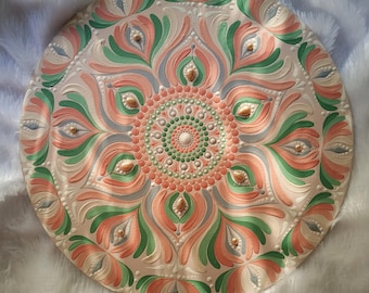 Può includere: Un piatto decorativo rotondo bianco sporco con un elaborato design a mandala. Il design presenta un cerchio centrale circondato da intricati motivi nei toni del corallo, verde, grigio e oro. Il piatto è probabilmente in ceramica o materiale simile.