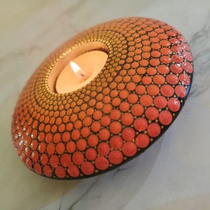 K&ouml;nnte beinhalten: Ein handbemalter, ovaler Kerzenhalter mit einem orangefarbenen und schwarzen Punktmuster. Eine brennende Kerze befindet sich in der Mitte. Der dekorative Artikel ist f&uuml;r ein Teelicht konzipiert.