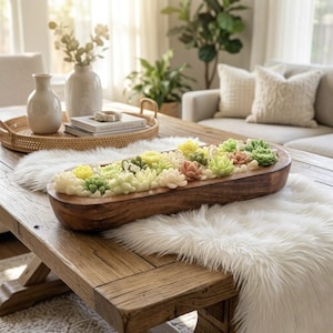 May include: A wooden trough filled with colourful succulent plants sits on a rustic wooden table. The succulents are various shades of green, yellow, and peach. A white faux fur rug and other decorative items are also on the table.