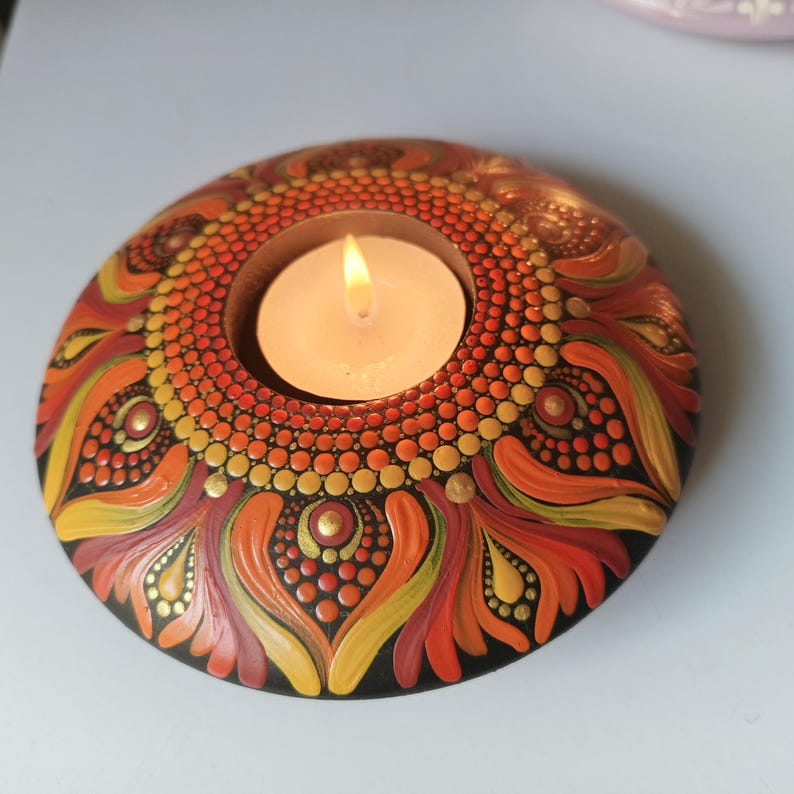 K&ouml;nnte beinhalten: Ein runder, handbemalter Kerzenhalter mit einer brennenden Kerze. Der Halter hat ein Mandala-Design in Orange-, Gelb- und Rott&ouml;nen mit goldenen Akzenten. Der Sockel ist schwarz und die Kerze ist wei&szlig;.