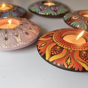 K&ouml;nnte beinhalten: Eine Sammlung handbemalter, dekorativer Kerzenhalter. Jeder Halter ist ein einzigartiges, ovalf&ouml;rmiges Design mit komplizierten Mustern und leuchtenden Farben, darunter Blau, Lila, Orange und Rosa. In jedem Halter befinden sich brennende Kerzen, die ein warmes Leuchten erzeugen.