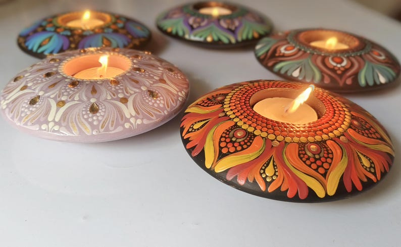 K&ouml;nnte beinhalten: Eine Sammlung handbemalter, dekorativer Kerzenhalter. Jeder Halter ist ein einzigartiges, ovalf&ouml;rmiges Design mit komplizierten Mustern und leuchtenden Farben, darunter Blau, Lila, Orange und Rosa. In jedem Halter befinden sich brennende Kerzen, die ein warmes Leuchten erzeugen.