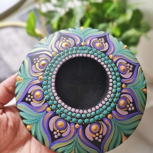 K&ouml;nnte beinhalten: Ein handbemaltes, rundes Dekorationsst&uuml;ck mit einer zentralen schwarzen Vertiefung. Das Design weist t&uuml;rkisfarbene, violette und goldene Akzente in einem Mandala-Stil auf. Das St&uuml;ck wird in einer Hand gehalten, wodurch die komplizierten Details und Farben hervorgehoben werden.