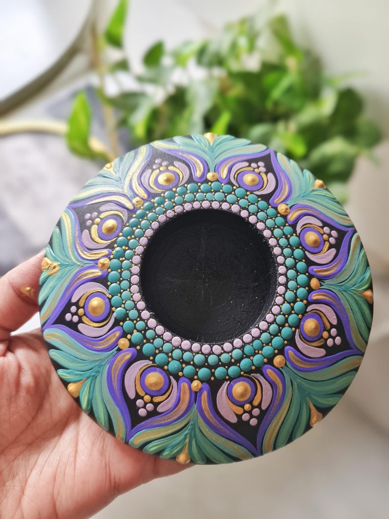 K&ouml;nnte beinhalten: Ein handbemaltes, rundes Dekorationsst&uuml;ck mit einer zentralen schwarzen Vertiefung. Das Design weist t&uuml;rkisfarbene, violette und goldene Akzente in einem Mandala-Stil auf. Das St&uuml;ck wird in einer Hand gehalten, wodurch die komplizierten Details und Farben hervorgehoben werden.