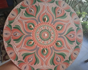 May include: A round, hand-painted decorative piece with a mandala design. The piece features a light pink background with a symmetrical pattern in shades of coral, green, and gray. Gold-colored accents are evenly spaced throughout the design.