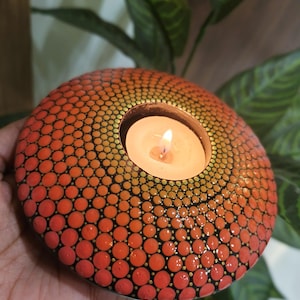 K&ouml;nnte beinhalten: Ein runder, orange-roter Kerzenhalter mit einem Punktmuster. Der Kerzenhalter hat eine brennende Kerze im Inneren. Der Hintergrund umfasst gr&uuml;ne Bl&auml;tter.