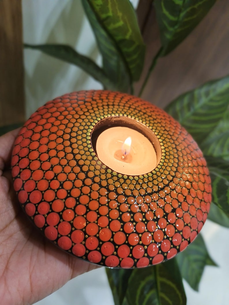 K&ouml;nnte beinhalten: Ein runder, orange-roter Kerzenhalter mit einem Punktmuster. Der Kerzenhalter hat eine brennende Kerze im Inneren. Der Hintergrund umfasst gr&uuml;ne Bl&auml;tter.