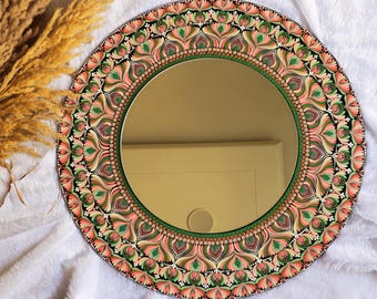 Può includere: Specchio decorativo rotondo con un motivo floreale dettagliato nei toni del rosa, verde e marrone su sfondo nero. Lo specchio ha un bordo verde e si trova su una superficie bianca.