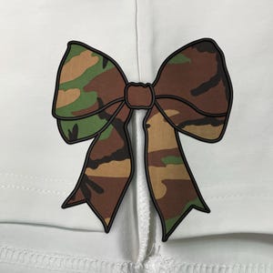 Könnte beinhalten: Ein weißes Hemd mit einer großen Tarnmuster-Schleife auf dem Rücken.