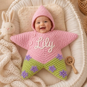 May include: A star-shaped crochet baby swaddle blanket with a hood, featuring a pink upper section with the name Lily embroidered in white script, two pink buttons, and a light green lower section decorated with purple flower appliques.
