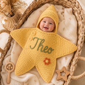 May include: A yellow star-shaped crochet baby swaddle blanket with a pointed hood. The garment features the name Theo embroidered in green yarn, two small buttons at the shoulder, and a small orange and yellow flower applique on the front surface.