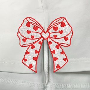 May include: A white fabric with a red glitter bow with small red hearts. The bow is centred on the fabric and has a white string attached to the centre.