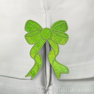 May include: A green glitter bow with a center loop is sewn onto a white fabric surface.