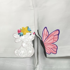 May include: White embroidered bunny with a floral crown, kissing a pink butterfly with purple accents on a white background.