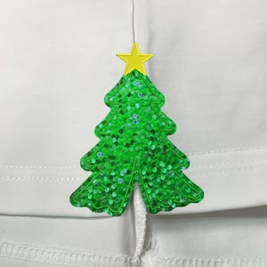 May include: A green Christmas tree applique with a yellow star on top. The tree is covered in green sequins and is attached to a white fabric. The tree is a festive decoration.