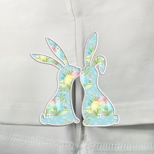May include: Two blue Easter bunnies with pastel floral and egg patterns kiss each other on a white background.