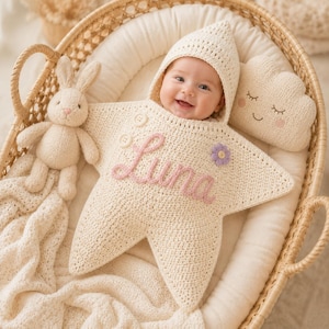 May include: A cream-colored crocheted star-shaped baby swaddle with a hood, featuring the name Luna embroidered in pink script, two white buttons, and a small purple flower accent. A plush rabbit toy and a cloud-shaped pillow are nearby.