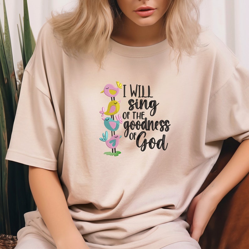 May include: A beige t-shirt with a colorful embroidered design of birds stacked on top of each other. The text "I will sing of the goodness of God" is embroidered in black below the birds.