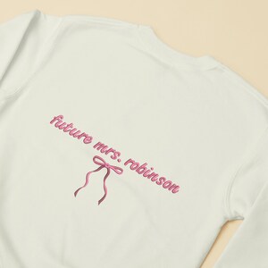 May include: White sweatshirt with the text "future mrs. robinson" in pink lettering with a pink bow.