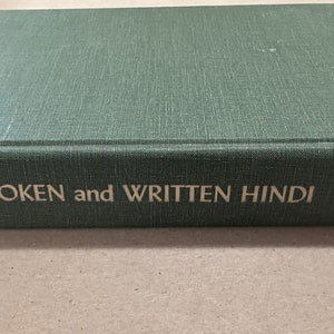 May include: A green hardcover book titled "Spoken and Written Hindi" by Fairbanks and Misra, published by Cornell University Press. The book is lying on a light brown surface.